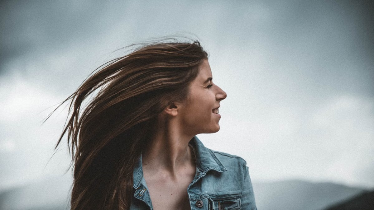 Portrait of a smiling young woman in a denim jacket against a cloudy sky.